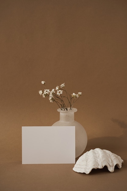 Tarjeta de hoja de papel en blanco con concha, hermosas flores blancas contra la pared marrón neutral.