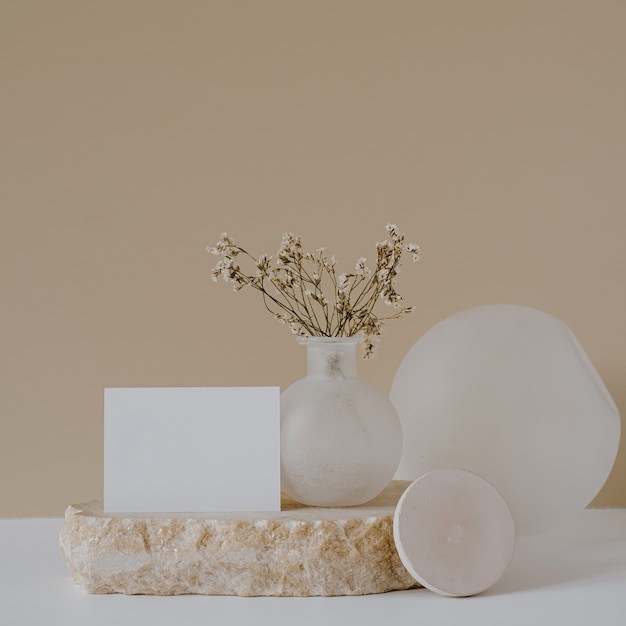 Tarjeta de hoja de papel en blanco con flores en piedra de mármol contra la pared beige neutra