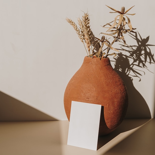 Tarjeta de hoja de papel en blanco con olla de barro con ramo de trigo seco con sombra de luz solar en blanco