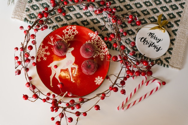 Tarjeta de Navidad. Placa roja con un ciervo, manzanas confitadas, dulces de caña y bayas rojas en las ramas sobre la mesa, vista superior