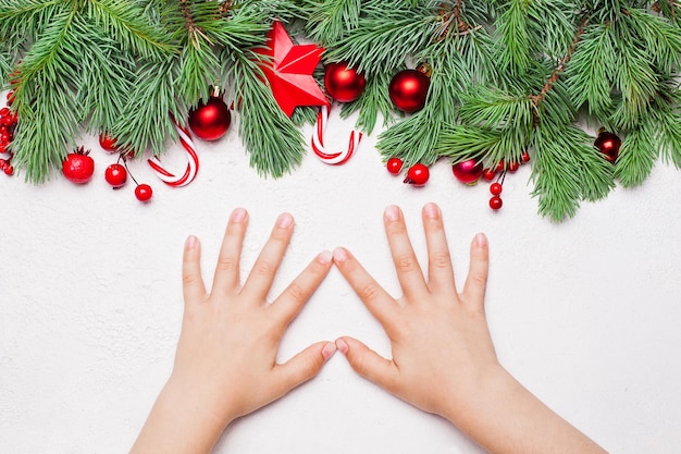 Tarjeta de Navidad con rama de abeto de Navidad verde adornos de bayas de acebo rojo y manos de niña sobre fondo blanco