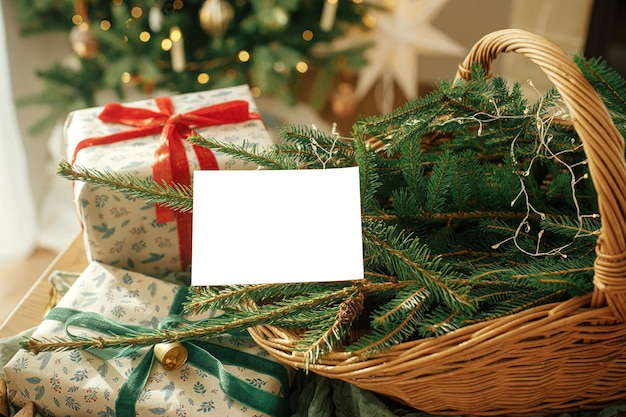 Tarjeta de Navidad simulada Tarjeta de felicitación vacía en el fondo de elegantes regalos envueltos en Navidad, ramas de abeto en una cesta rústica y árbol festivo con luces doradas Espacio para texto