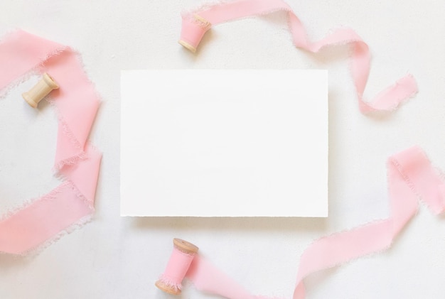 Tarjeta de papel sobre mesa de mármol blanco con flores rosadas secas y vista superior de cintas de seda. Endecha plana con tarjeta en blanco horizontal. Maqueta de invitación romántica o tarjeta de felicitación, espacio de copia