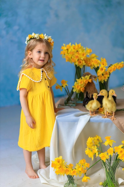 Tarjeta de pascua hermosa linda chica con un vestido amarillo brillante se sienta junto a conejos y patitos cerca de ramos de narcisos