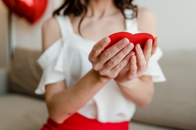 Tarjeta de San Valentín roja en forma de corazón en manos de mujer en el sofá en casa