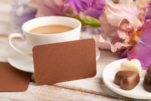 tarjeta de visita marrón con taza de cioffee, caramelos de chocolate y flores de iris sobre superficie blanca.