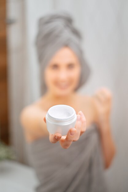 Foto tarro blanco con una crema para las arrugas e hidratante de la piel en manos de una chica difuminada en el baño.