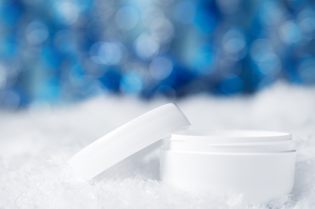 Foto tarro de crema blanca sobre la nieve con un fondo brillante borroso. la decoración de navidad borrosa bokeh brilla en azul. cuidado de la piel seca en invierno. producto de belleza cosmético para el cuidado de la piel del rostro y las manos.