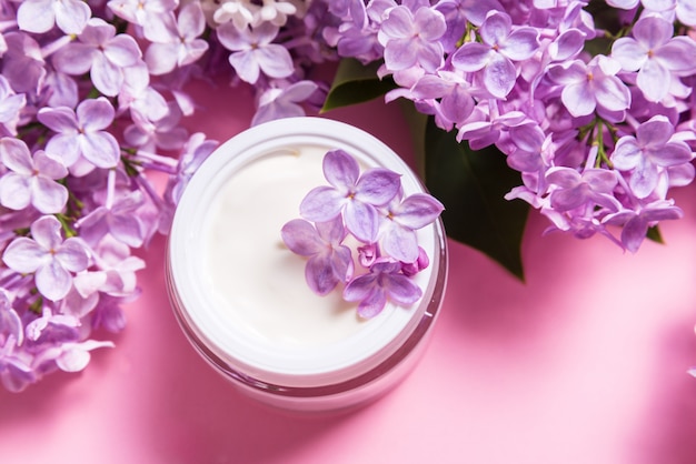 Tarro de crema facial natural con flores de color lila sobre fondo rosa