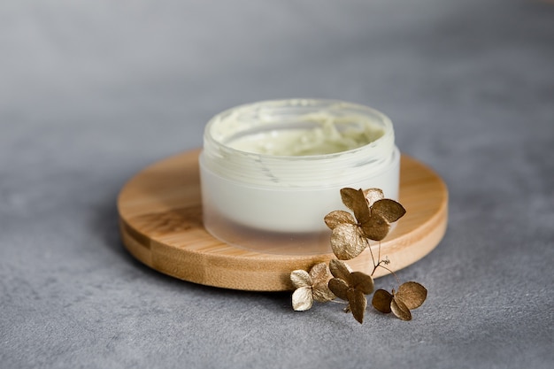 Un tarro de crema facial sobre un soporte de madera sobre una mesa gris.