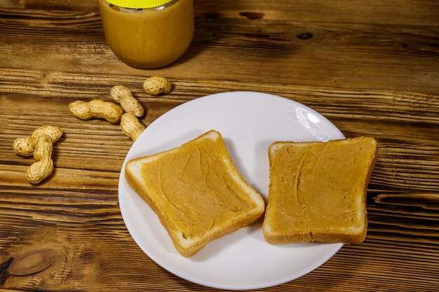 Tarro de mantequilla de maní y plato con dos sándwiches con mantequilla de maní en mesa de madera