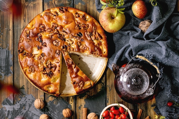 Tarta casera de manzana y nueces Charlotte en un plato con manzanas frescas de jardinería, nueces, té de rosa mosqueta alrededor sobre fondo de tablones de madera oscura. Beber té otoñal. Vista superior