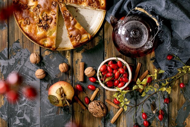 Tarta casera de manzana y nueces Charlotte en un plato con manzanas frescas de jardinería, nueces, té de rosa mosqueta alrededor sobre fondo de tablones de madera oscura. Hornear en casa otoñal. Endecha plana