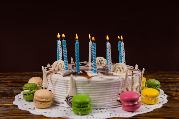 Tarta de cumpleaños casera blanca con un montón de velas encendidas cerca de macarons de diferentes colores, sobre un escritorio de madera