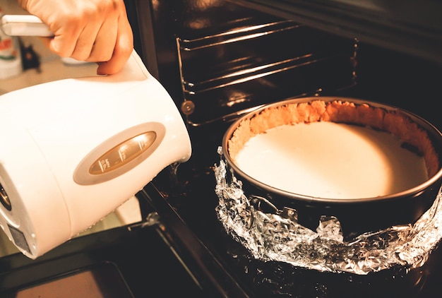 Tarta de queso en el horno. Forme un molde desmontable en papel de aluminio. La mujer está vertiendo agua de la tetera. Proceso de horneado. Postres caseros. Pastelería sabrosa.