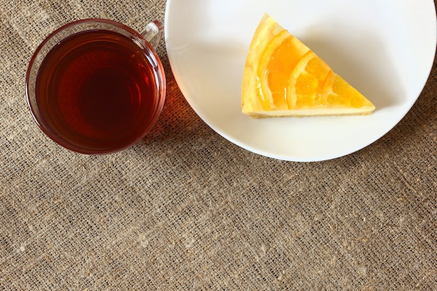 Tarta de queso con naranjas en un plato blanco, taza de vidrio con té sobre mantel de arpillera. Vista superior.