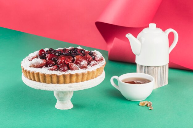 Tarta de requesón con bayas sobre un soporte de madera sobre un fondo multicolor con una tetera y una taza de té