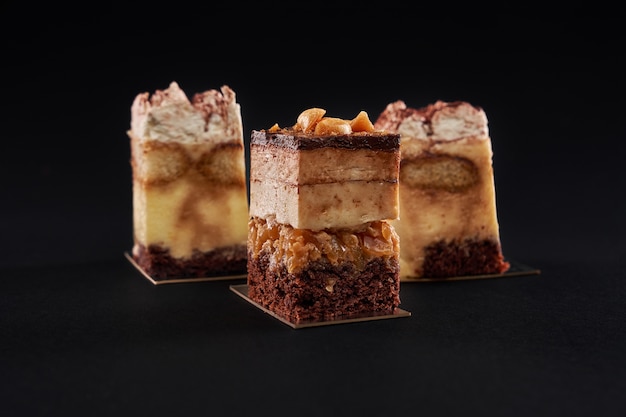 Tarta de tiramisú con tres capas de galleta de chocolate y sirope de café natural con coñac y nata. Tres piezas cuadradas en fila decoradas con crema batida y cacao, aisladas en pared negra.