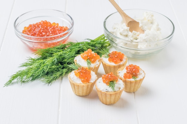 Tartaletas con caviar rojo y un recipiente de vidrio con requesón sobre una mesa de madera blanca. Aperitivo con delicias del mar.