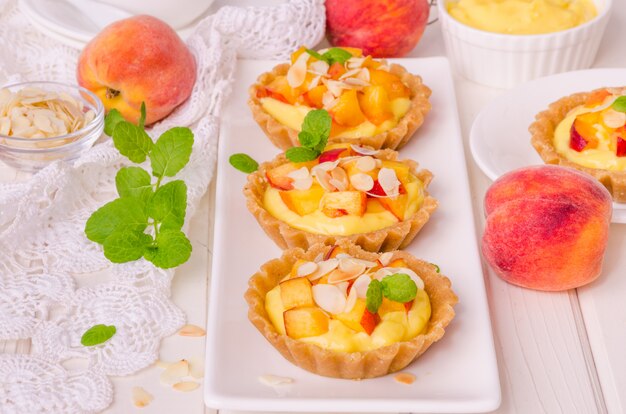 Tartaletas con crema pastelera, duraznos frescos y almendras sobre fondo blanco de madera