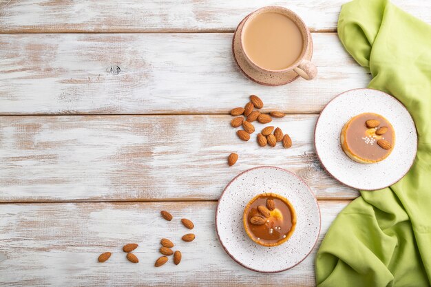 Tartaletas dulces con crema de almendras y caramelo con taza de café sobre un fondo de madera blanca y textil verde. vista superior, endecha plana, espacio de copia.