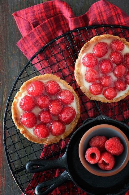 Tartaletas de frambuesa roja con crema de vainilla y frambuesas frescas glaseadas sobre una rejilla para enfriar sobre madera marrón oscuro