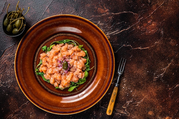 Tartar o tartar con pescado de salmón, cebolla morada, rúcula y alcaparras en plato rústico. Fondo oscuro. Vista superior. Copie el espacio.