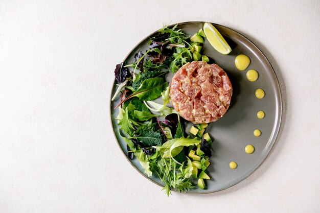 Tártaro de atum com salada verde, limão, abacate e molho de mostarda servindo em prato de cerâmica sobre mesa de textura branca. Postura plana, copie o espaço. Jantar fino, aperitivo de restaurante