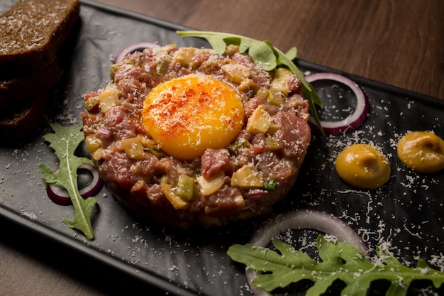 Tártaro de carne com mostarda de ovo de cebolas verdes e pão em uma placa preta Vista superior de tártaro