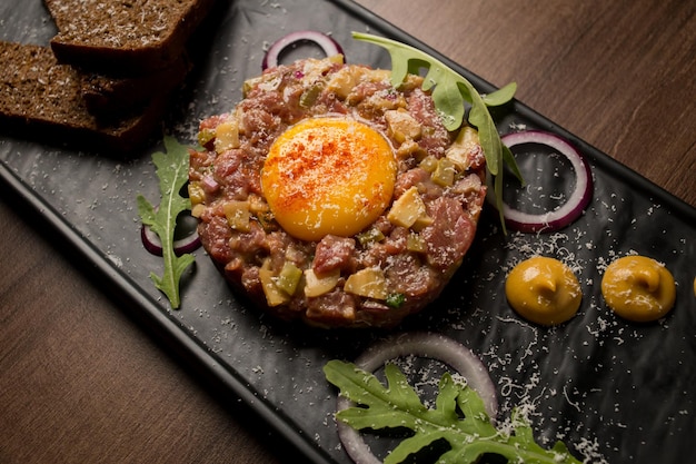 Tártaro de carne com mostarda de ovo de cebolas verdes e pão em uma placa preta Vista superior de tártaro