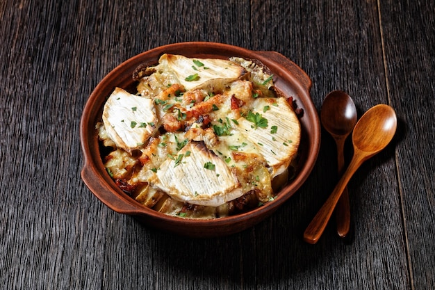 Tartiflette, batata francesa, bacon e caçarola de queijo em um prato de barro em uma mesa de madeira escura com colheres, cozinha francesa