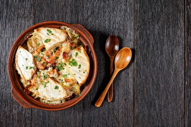 Foto tartiflette, cazuela de patatas, tocino y queso francés en un plato de barro sobre una mesa de madera oscura con cucharas, plano, espacio libre, cocina francesa