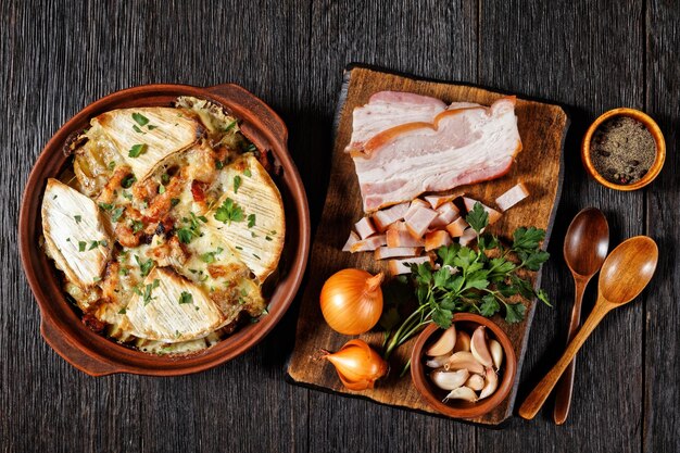 Tartiflette, patata francesa, tocino y cazuela de queso en una cazuela de barro con ingredientes sobre una tabla para cortar, plano, cocina francesa