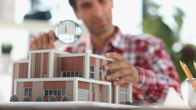 Tasador masculino mirando el modelo de la casa a través de la evaluación de bienes raíces con lupa y la búsqueda de la casa