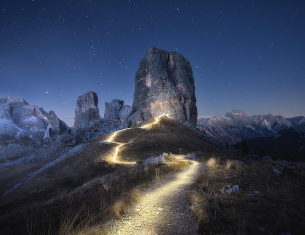 Taschenlampenpfade auf einem Bergpfad vor hohen Felsen in der Nacht im Herbst. Dolomiten, Italien. Bunte Landschaft mit Lichtpfaden, Pfad auf dem Hügel, alpine Berggipfel, Himmel mit Sternen im Herbst