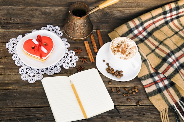 Tasse Cappuccino, herzförmige Kekse breite Nachricht, Notizbuch, Bleistift und Kaffeekannen auf braunem Holztisch