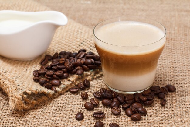 Tasse Cappuccino, Sahnesauce, Kaffeebohnen und Leinensack auf Sackleinen.