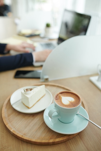 Tasse Cappuccino und Kuchen auf dem Café-Tisch der Geschäftsfrau, die am Laptop arbeitet