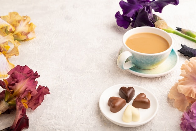 Tasse Cioffee mit Pralinen und lila und burgunderroten Irisblumen auf grauem Hintergrund.