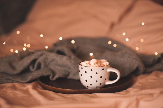 Tasse heiße Schokolade mit Marshmallow mit Strickstoff auf Holztablett über Weihnachtsbeleuchtung im Bett Nahaufnahme Winterferienzeit