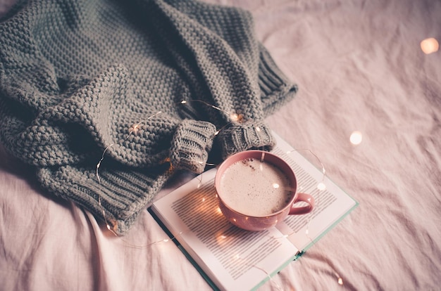 Tasse heißen Kaffee bleiben auf offenem Buch mit Strickpullover im Bett Nahaufnahme mit Lichtern im Hintergrund. Guten Morgen. Winterferienzeit.