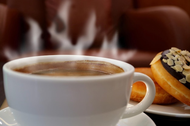 Tasse heißen Kaffee mit aufsteigendem Dampf und einem Teller Donuts im Hintergrund