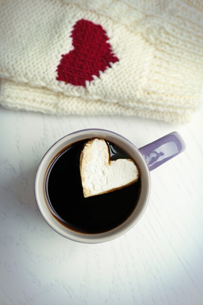 Tasse heißen Kaffee mit Herz Marshmallow und warmen Handschuhen auf hellem Holztisch, Nahaufnahme