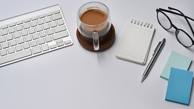 Tasse heißen Kaffee, Notizblock, Haftnotizen und Brillen auf weißem Schreibtisch.