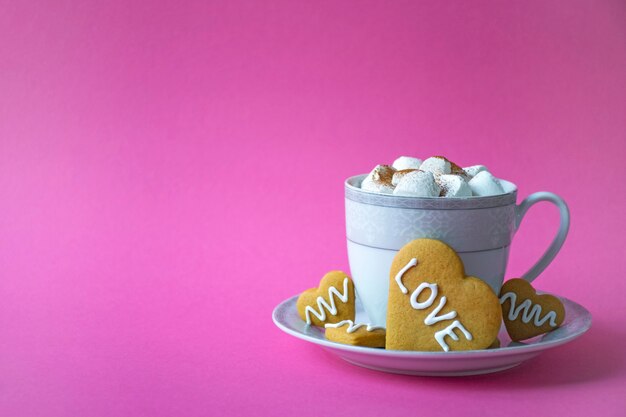 Tasse heißen Kaffee oder Kakao mit Marshmallow und Keks in Herzform mit Liebesphrase auf Rosa