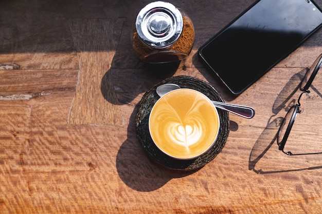 Tasse heißen Milchkaffee mit Latte Art, Smartphone, Brillen und braunem Zucker