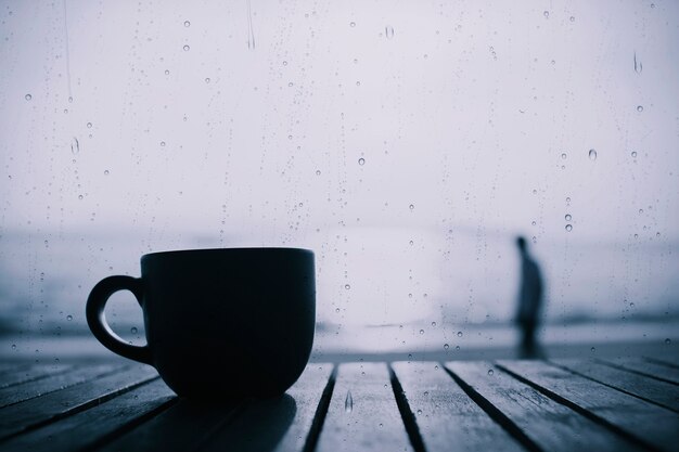 Tasse Kaffee auf Holztisch morgens mit Regen fällt auf Glas