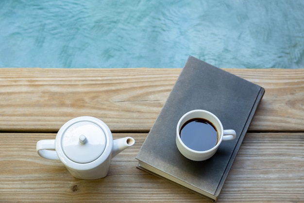 Tasse Kaffee auf Lesebuch mit Kaffeekanne auf Bretterboden durch Swimmingpool.