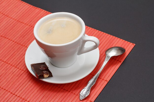 Tasse Kaffee auf Untertasse mit Stück Schokoriegel, Löffel und Bambusserviette auf schwarzem Hintergrund. Ansicht von oben.