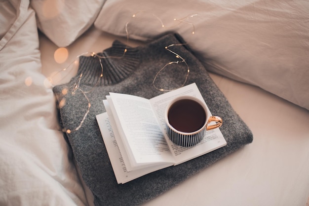 Tasse Kaffee bleibt auf gestricktem Textilpullover im Bett mit offenem Papierbuch mit weihnachtlich leuchtenden Lichtern aus nächster Nähe